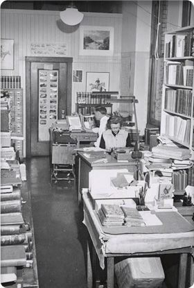 View of Richard Dillon and an unidentified staff member seated at desks in the Sutro Library Research Room. Books stacked on the tables and on shelves; bust of Adolph Sutro at left.