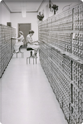 Union card file lining on both sides of corridor. Two women at work filing.